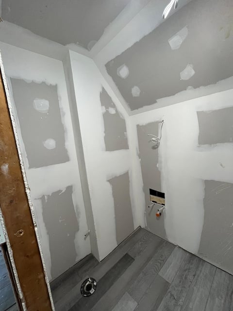 Interior room under construction with drywall sheets, exposed studs, and patch holes during renovation