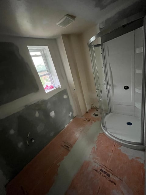 Unfinished bathroom under renovation with exposed drywall, corner shower enclosure, small window, and bare flooring