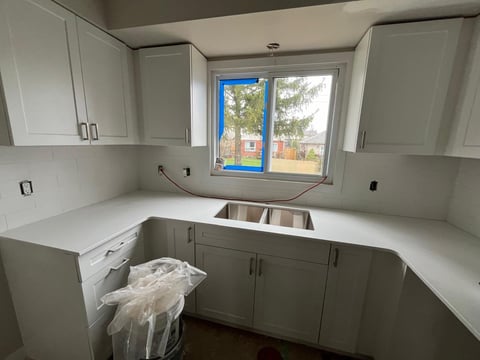 Modern kitchen under renovation with white cabinets, stainless steel sink, and white countertops, featuring a window with blue frame overlooking a yard