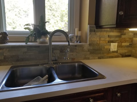 Modern kitchen sink with double basin, stainless steel faucet, and tile backsplash. Countertop with green plant and window visible above