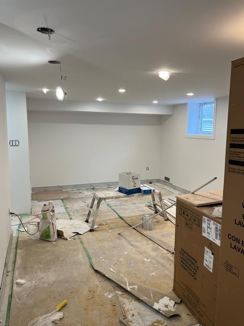 Unfinished basement room under construction with exposed flooring, drop cloths, ladders, and recessed ceiling lights installed