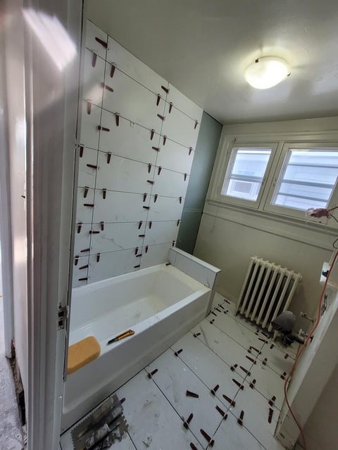 Bathroom under renovation with white bathtub, geometric tile pattern, ceiling light, radiator, and windows with blinds