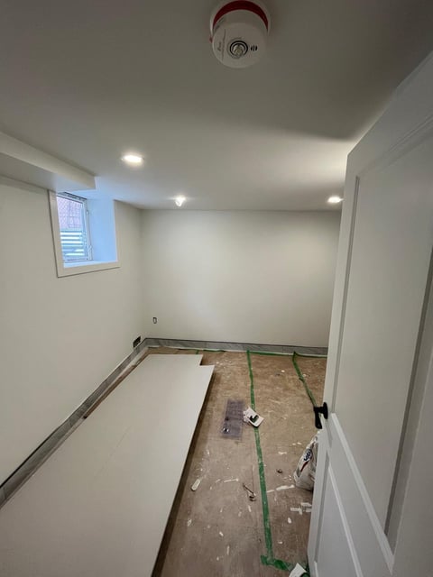 Empty basement room under construction with white drywall, recessed lighting, a small window, and concrete flooring with green marking tape